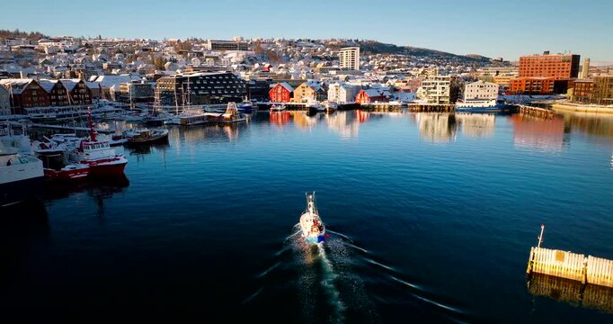 Drone view following fishing boat sailing in blue water leaving wake in frozen harbor in Tromso, Norway. Aerial forward