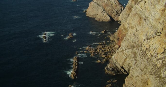 Handheld shot captures rugged Cabo Pe&ntilde;as cliffs with native marine plants, waves hitting rocks, and serene Atlantic seascape on the Asturias coastline under clear natural light.