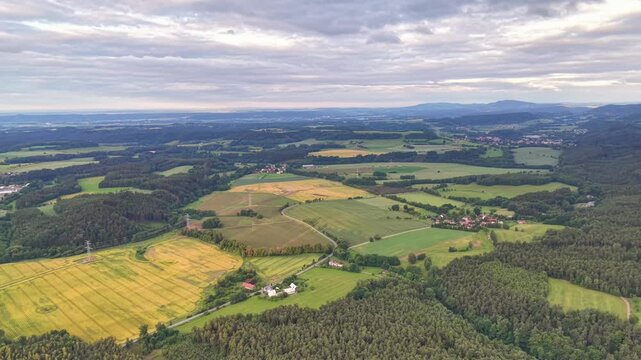 Broad aerial panorama of patchwork fields and meadows across rolling hills, with forest patches, winding roads, scattered villages, power lines, orchards, windmills, and cloudy sky