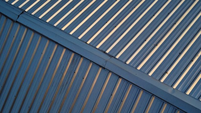Architectural detail of a blue corrugated metal roof featuring a ridge cap, geometric diagonal patterns, and contrasting sunlight shadows