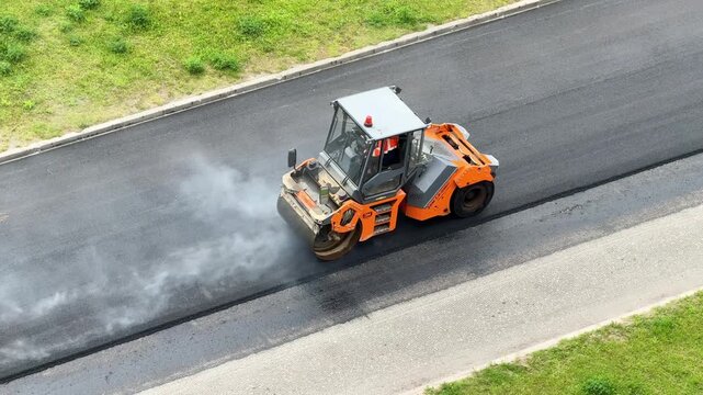 Orange road roller compacts a newly paved highway, transport infrastructure improvement, civil engineering, industrial maintenance, aerial roadwork scene.