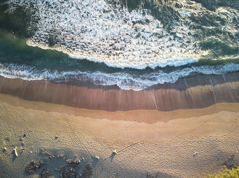 Aerial view of a sandy beach with gentle waves washing onto the shore, featuring visible footprints in the sand and soft golden light in Jinotepe, Carazo, Nicaragua.