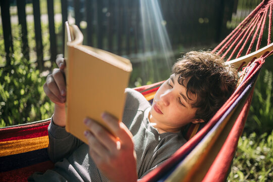 A boy reads a book while relaxing in a backyard hammock