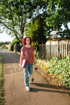 Teenage girl student walks with a skateboard and folder on sidewalk