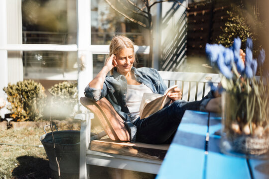 Mature woman reading book sitting on bench at front yard