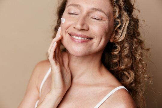 A mature woman with natural skin smiles softly while applying cream, conveying authentic skincare and calm beauty in a warm, candid studio moment.
