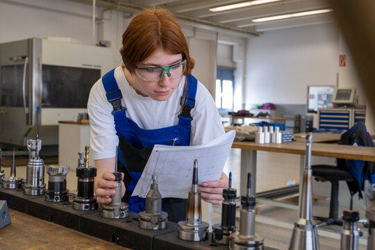 Female technician working with metallic machinery parts while reading book at workshop