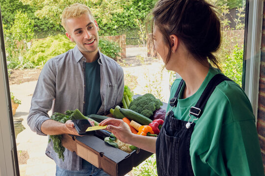 Woman paying and talking to delivery person with fresh vegetables near doorway