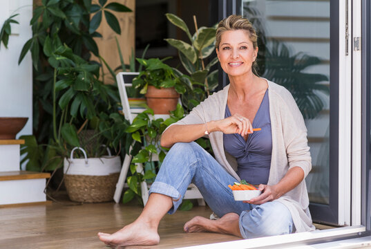 Portrait of smiling mature woman sitting on the floor at open terrace door eating finger food
