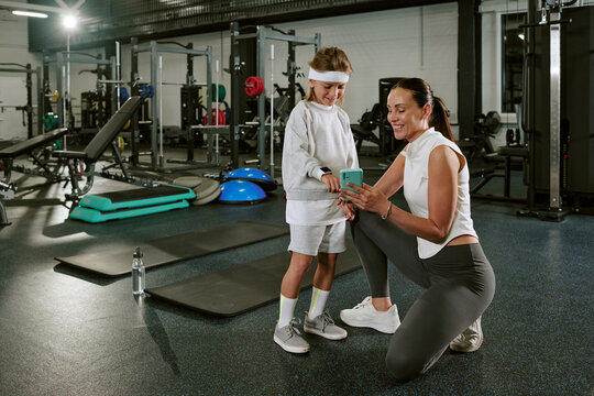 Caucasian female trainer showing smartphone to Caucasian child in gym, tracking fitness progress. Image suitable for promoting youth sports programs and digital health tracking apps