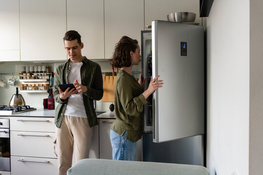 Young man using tablet PC by woman opening refrigerator in kitchen at home