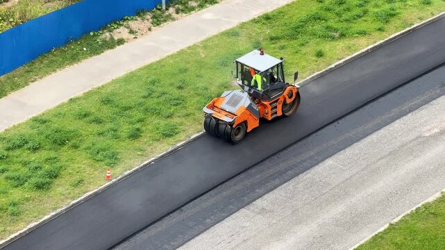 Orange road roller compacts fresh asphalt on a highway, transport infrastructure improvement, civil engineering, industrial road maintenance, aerial construction view.