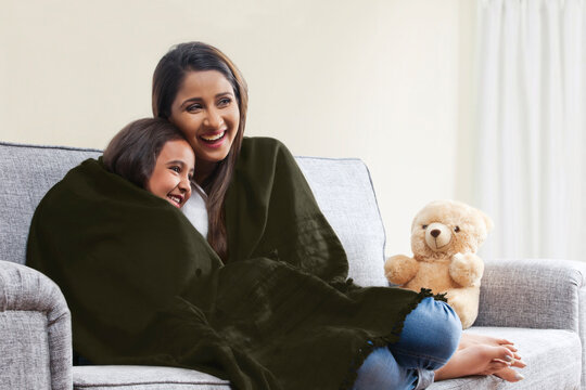 Smiling mother and daughter wrapped in blanket watching TV on living room sofa