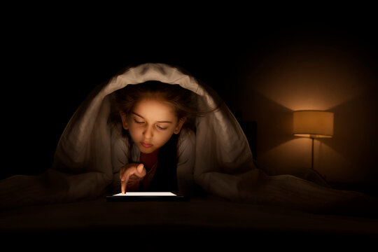 Young girl using digital tablet under bed sheet at night in bedroom