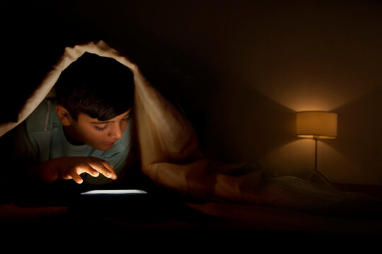 Young boy using digital tablet under blanket at night in bedroom