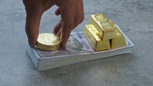 Hand places gold bitcoin coins on stack of US dollar bills next to gold bars for showing wealth and assets