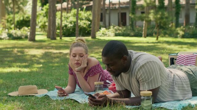 Bored interracial couple using smartphones during park picnic