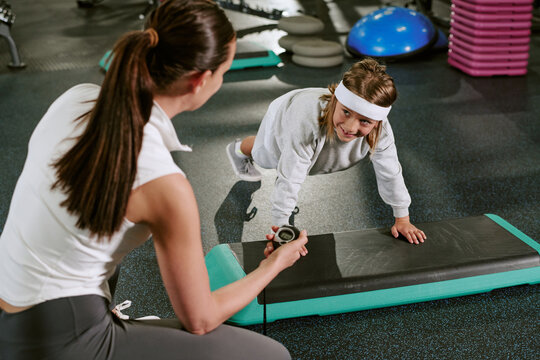 Caucasian female trainer timing Caucasian child doing plank exercise on step platform in gym. Image suitable for promoting youth fitness programs and children sports coaching