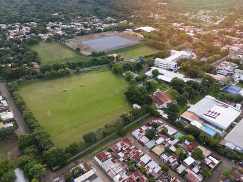 Aerial view of a sports complex featuring a large green soccer field, an adjacent stadium with a track, and surrounding residential buildings in San Marcos, Carazo, Nicaragua.