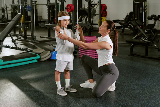 Caucasian female trainer helping Caucasian child lifting dumbbells in gym, promoting youth fitness. Image suitable for children sports programs and healthy lifestyle marketing