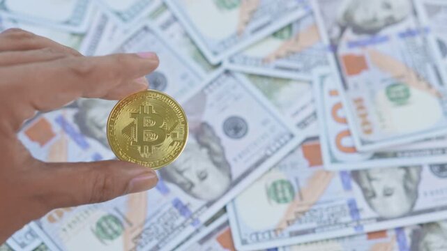 Hand holds gold bitcoin coin against background of scattered US dollar bills for depicting crypto vs fiat currency