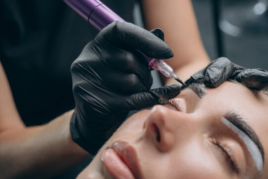 Permanent makeup artist performs an eyebrow tattoo procedure after mapping the shape, creating natural brows with a PMU machine in a professional beauty studio.