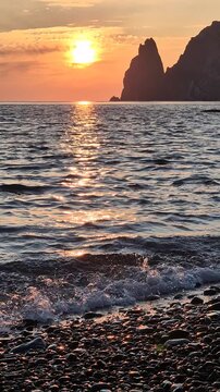 Golden sunset ocean waves wash over a rocky pebble beach with towering coastal cliffs. Static wide angle shot.