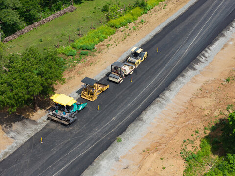 Aerial view of road construction with paving machinery, including a paver and rollers, on a newly laid asphalt road surrounded by green trees in Jinotepe, Carazo, Nicaragua.