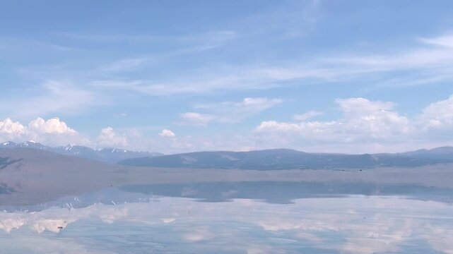 Witness the breathtaking mirror image of a pristine blue sky and fluffy clouds on the calm surface of Khuvsgul Lake in Mongolia. Distant snow-capped mountains add to this tranquil, majestic vista.