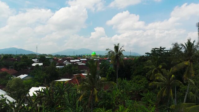 Green mosque dome rises above dense tall palm trees and red tile rooftops in Pelowok Selatan, Kediri, West Lombok Regency, misty mountain ridges spread beyond village. Aerial drone rising shot