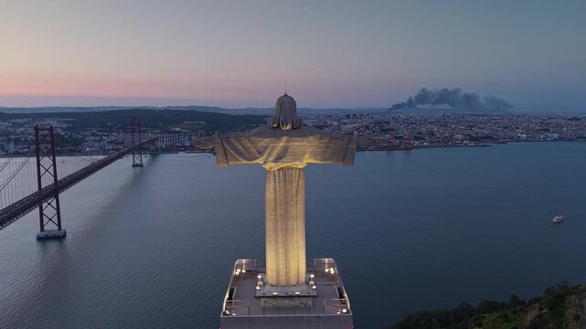 Aerial twilight view behind the illuminated Cristo Rei monument toward the 25 de Abril Bridge and Lisbon, with dark smoke rising above the city across the Tagus River.