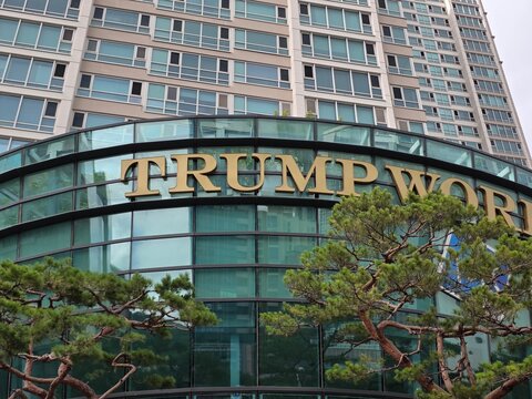 July 8th 2026,Low angle shot of the Trump World building exterior facade with large golden letters, glass curved structure, and green pine trees in the foreground.