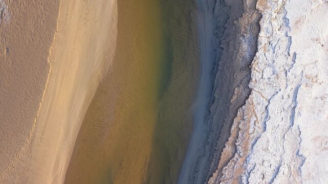 Aerial view reveals river flowing through sandy banks. Water glows greenish-yellow. Salt flats display white crystalline patterns. River moves through varied terrain. Friskney Flats