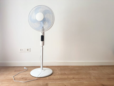 A white pedestal fan by a wall with its cord unplugged on a wooden floor. Electric fan for cooling a home in summer
