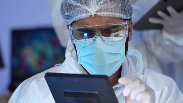 Scientist in hazmat suit safety goggle hairnet surgical mask operating microscope computer while conducting experiment inside high tech research laboratory