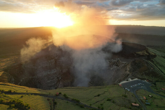 Aerial view of Masaya Volcano with volcanic gas and smoke rising from the crater at sunset, showing the rugged landscape and parking area in Nindiri, Masaya, Nicaragua.