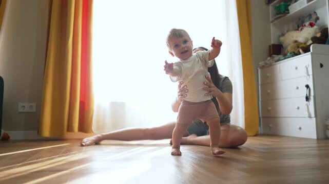 Toddler learns to walk. Mother supports toddler taking first step indoors. Toddler makes confident step forward. Precious bonding moment as mother guides toddler to walk. step walk journey with mother
