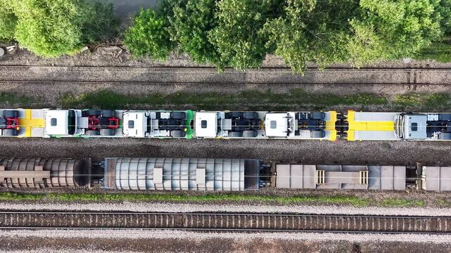 New trucks being transported on railway platforms, showcasing a continuous flow of vehicles on flatbed cars, with surrounding greenery and railway tracks visible in the background. Aerial view