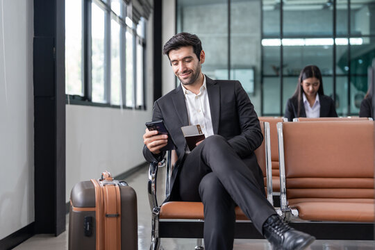 young male executive in suit holding passport with boarding pass, sitting in modern airport terminal checking time on a smartphone,preparing for corporate business trip or international flight