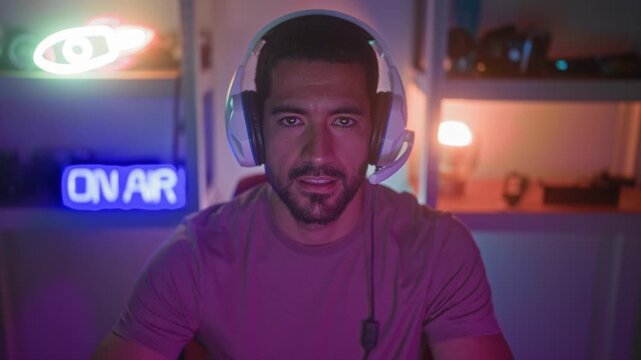 Man with beard wearing headset and microphone leans forward holding a gamepad, mouth slightly open and eyes fixed, neon on air sign and shelves visible in studio; focused concentration.