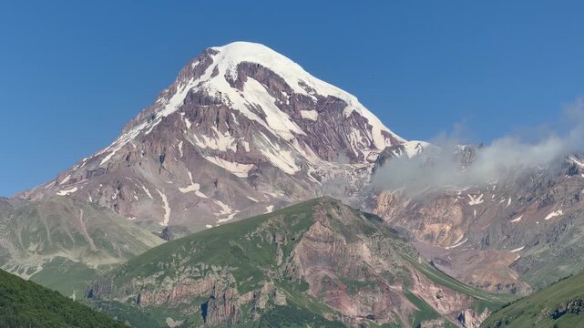 Majestic snow-capped Mount Kazbek peak soaring into clear blue sky above lush green hills in Kazbegi, Georgia. Spectacular natural landscape of Caucasus mountains under bright summer daylight.