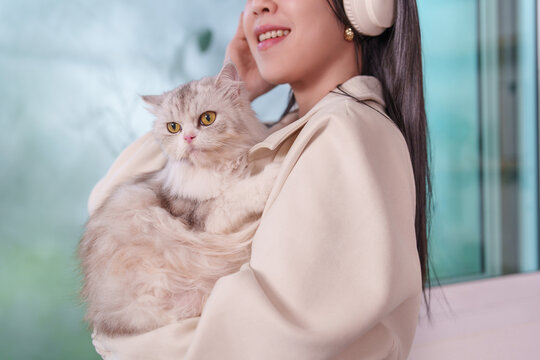 Happy Asian woman wearing headphones holding cute fluffy cat at home