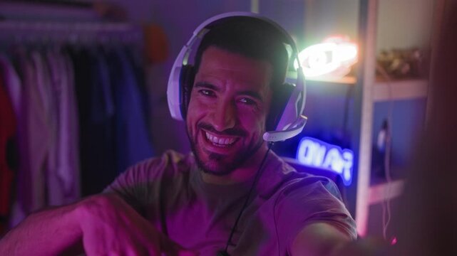 Man wearing gaming headset and mic, smiling, waving and pointing at camera while seated in studio, neon signs and clothing rack visible; joy community.