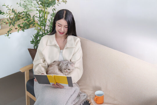Asian woman reading book while relaxing with cat on sofa at home