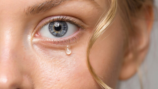 Macro of a young woman with blue eyes and blonde hair with a tear drop falling from her lower eyelid on a neutral background