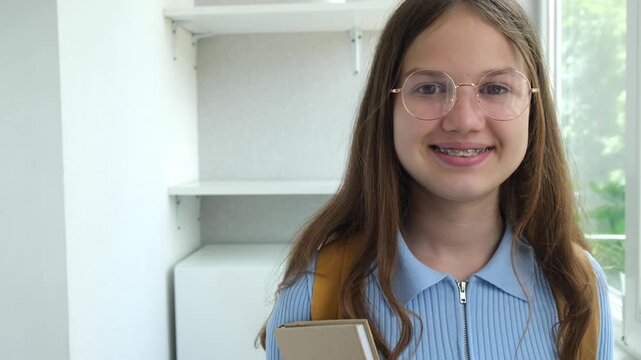 Portrait of smiling teenage student girl with book at home, copy space. Happy female teen in glasses with dental braces looking at camera