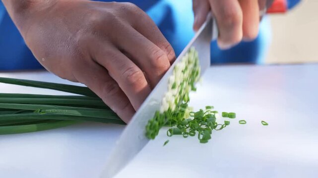 A culinary professional finely slices vibrant green scallions on a clean white cutting board before combining the fresh herbs with raw minced meat inside a polished metal bowl for meal preparation