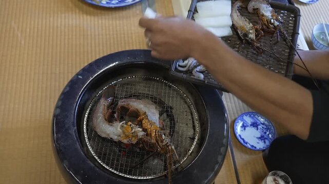 Hands place raw lobster and squid pieces onto a hot metal wire mesh grill over glowing coals, turning the seafood until cooked and peeling open the savory roasted meat