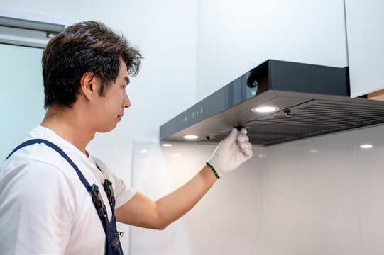 Asian male maintenance worker repairing modern cooker hood in domestic kitchen. Technician man wearing gloves cleaning kitchen range hood filter for clear ventilation. Home appliance service on site