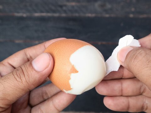 Close up view of human hands peeling the shell of a hard boiled brown chicken egg over a dark wooden background. Concept of healthy breakfast preparation, cooking, protein food, and kitchen routine